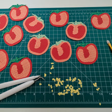 Load image into Gallery viewer, A collection of handmade paper tomato cutouts in red, pink, and green, arranged on a green cutting mat alongside crafting tools.