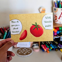 Load image into Gallery viewer, A hand holds a yellow kitchen-themed card with illustrations of a tomato and speech bubbles reading 'How do you fix a sliced tomato?' and 'With tomato paste!'. The background shows a blurred desk with colorful pens, markers, and art supplies.
