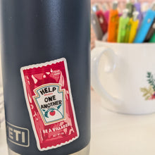 Load image into Gallery viewer, A close-up of a dark navy Yeti travel tumbler with a humorous sticker that says 'HELP ME ONE ANOTHER BE A VILLAGEAN' in a ketchup-pack design. A white ceramic mug filled with colorful pens is blurred in the background.

