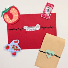 Load image into Gallery viewer, A flat-lay of handmade Valentine's Day themed stationery, featuring a red envelope with a 'Everyday Envelope' sticker, a kraft paper pouch with a strawberry heart charm, a strawberry-shaped sticker, and a small pink candy packet on a white background.

