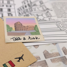 Load image into Gallery viewer, A small illustrated postcard featuring the Colosseum in Rome with the handwritten phrase 'Take a risk' on it, styled on top of printed travel-themed sheets and a kraft envelope with an Italian flag and airplane stamp.
